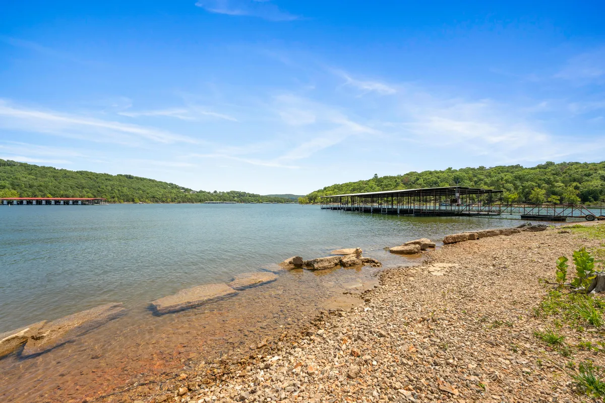 A sweeping view of Table Rock Lake with docks stretching over the water, green hills in the background, and a rocky shoreline. The tranquil water reflects the sky, and the scene evokes a sense of peaceful escape and natural beauty.
