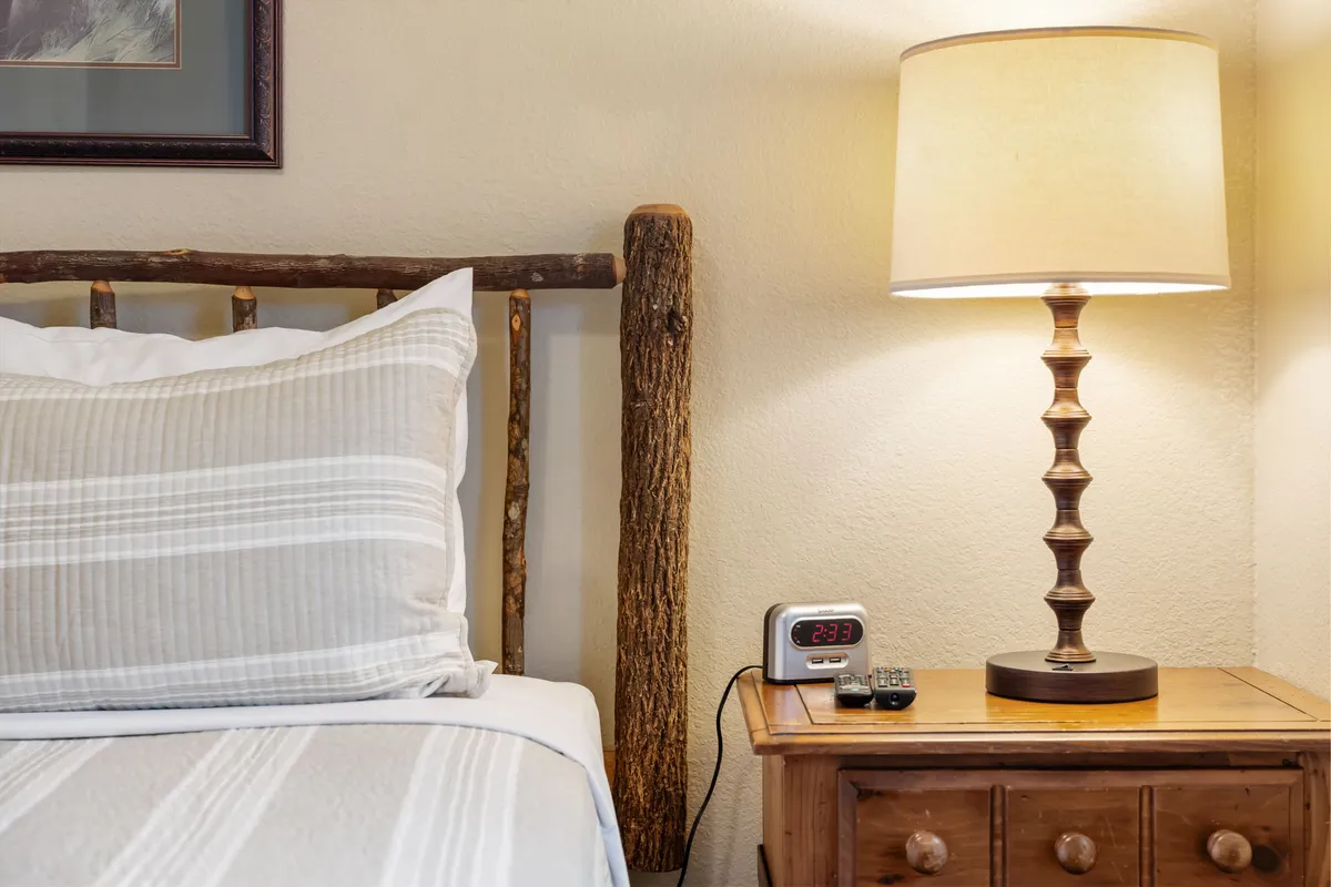 A rustic wooden queen bed frame adorned with striped pillows, a warmly lit table lamp, and a nightstand with a clock, all set against a soft-toned backdrop. The room evokes a sense of warmth, comfort, and intimacy perfect for restful slumber.