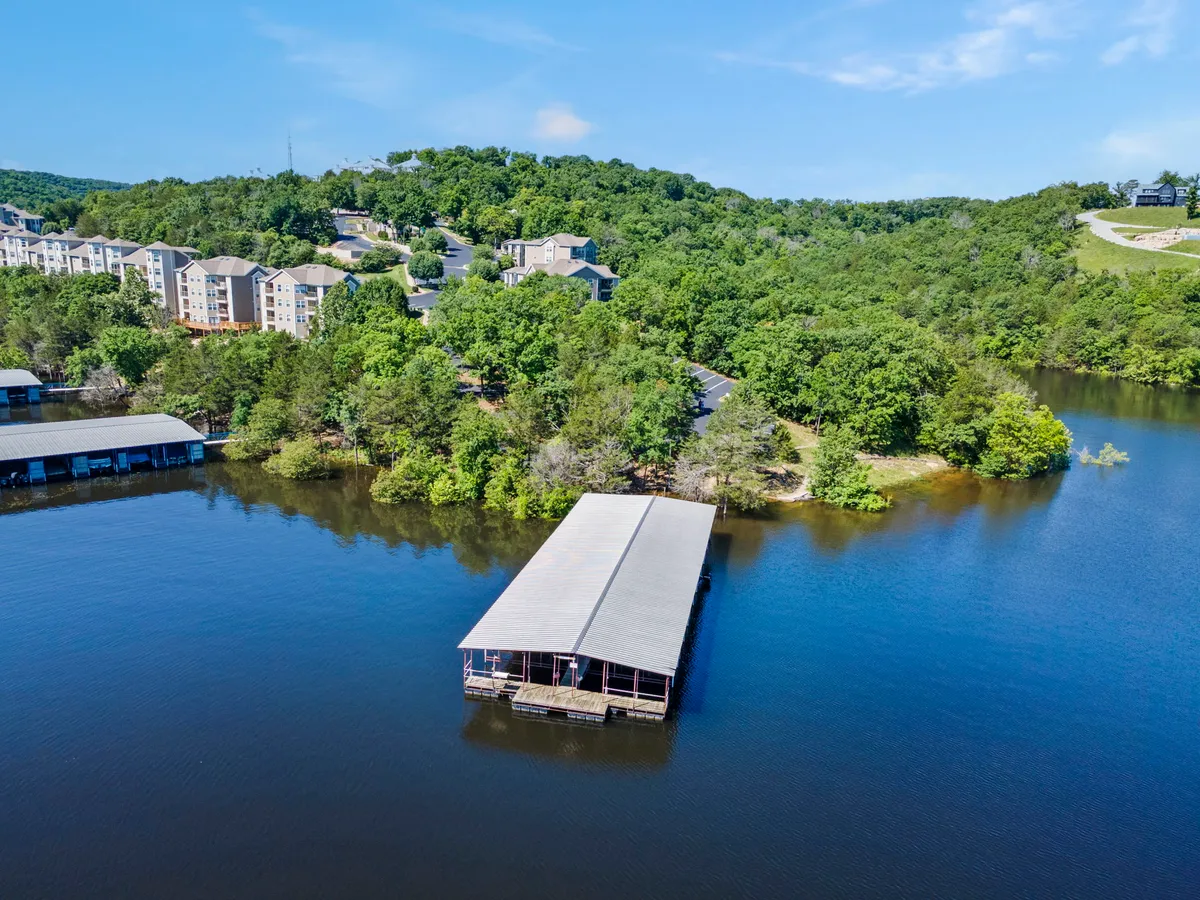 A tranquil dock stretches out over calm water, surrounded by lush green landscape and with residential buildings visible in the background. The scene captures the serenity of direct lake access and the perfect spot for fishing or launching a boat.