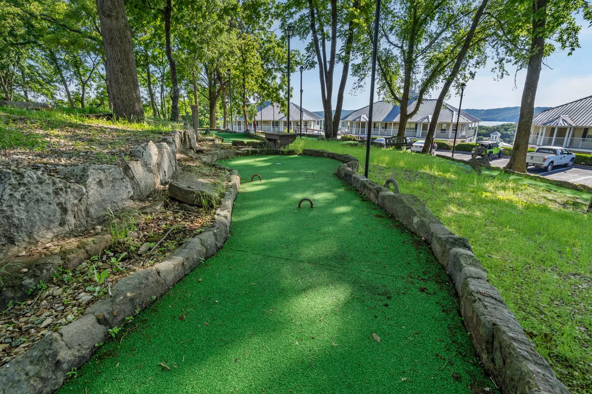 The photo showcases a well-maintained mini-golf course surrounded by green artificial turf, mature trees, and rock borders. The scene is bright and lively, inviting friendly competition. The course is well-suited for socializing and fun group activities in a natural outdoor setting.