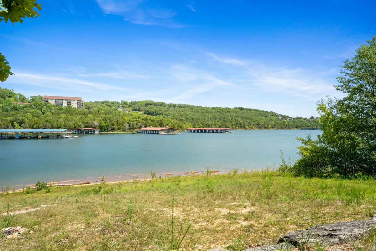 A wide view of a calm lake bordered by lush greenery and dotted with boathouses and distant hills. The serene water and tree-lined shore suggest opportunities for outdoor adventure, wildlife observation, and relaxed exploration.