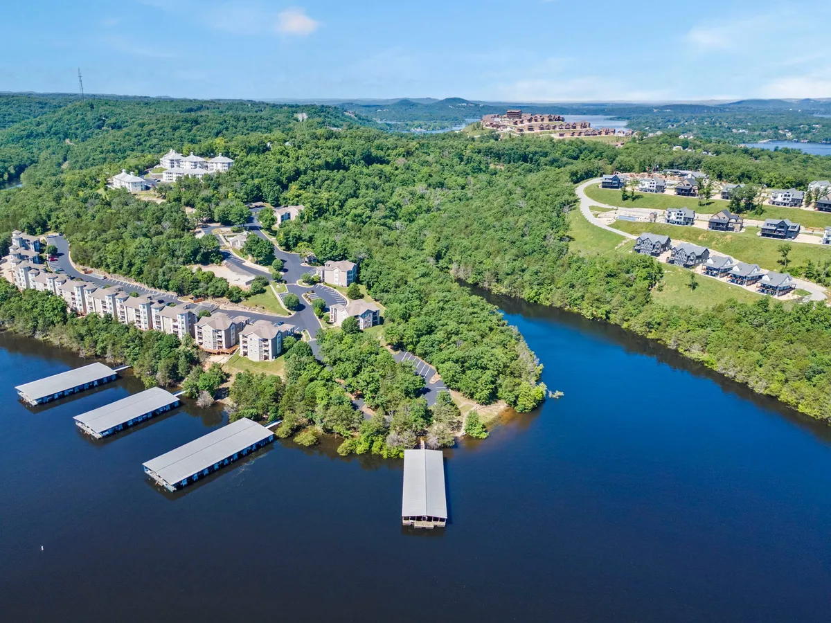 This exterior image features lush green hills, a serene waterfront with docks, residential buildings nestled in the landscape, and gently curving roadways. The environment is tranquil and inviting, suggesting a destination perfect for outdoor gatherings and explorations. The scene is sun-dappled and exudes the mood of a peaceful lakeside retreat.