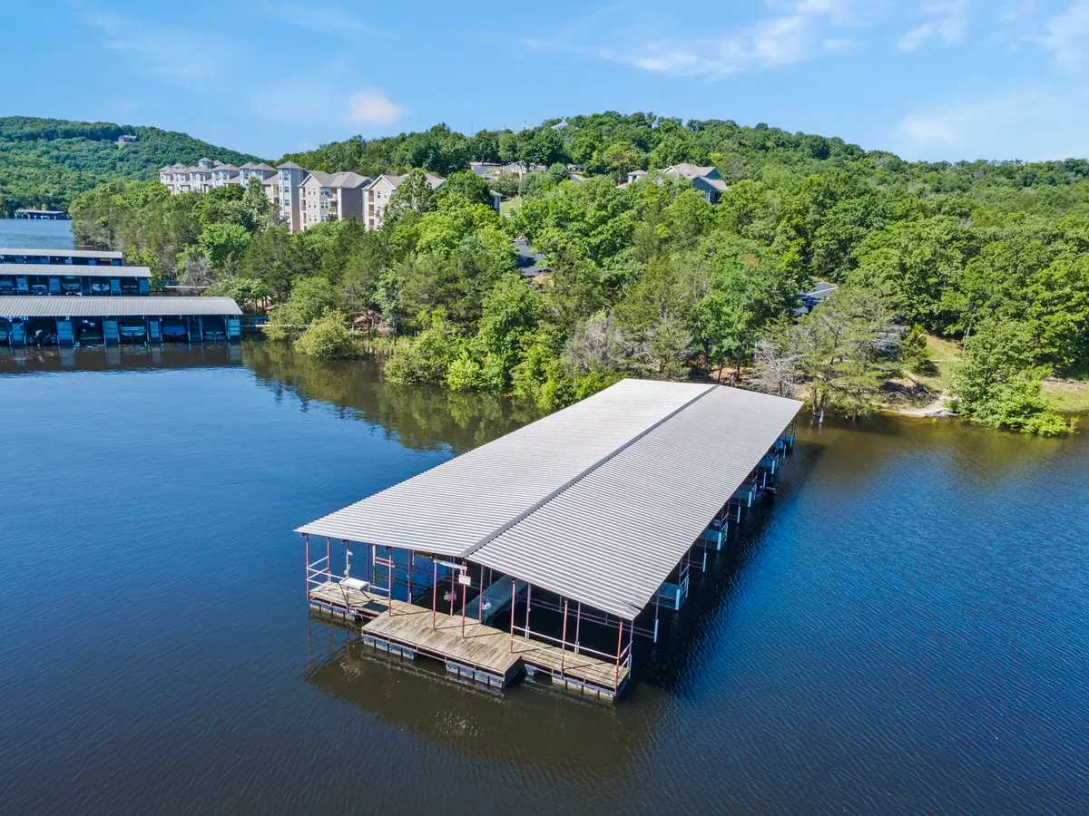 A tranquil lakeside exterior showing a boat dock, lush greenery, and nearby residential buildings. The walkway to the dock is visible, evoking the convenience and beauty of lakefront living.