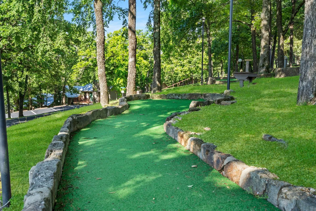 An exterior scene showing a putting green surrounded by trees, a stone pathway, and park benches. The space is playful and relaxed, suggesting outdoor activities like mini-golf, picnicking, and family fun.