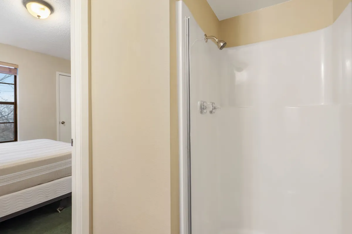 A contemporary ensuite bathroom featuring a glass shower stall, visible bedroom window, and part of the bed. The clean lines and soft lighting create a sense of privacy and rejuvenation.