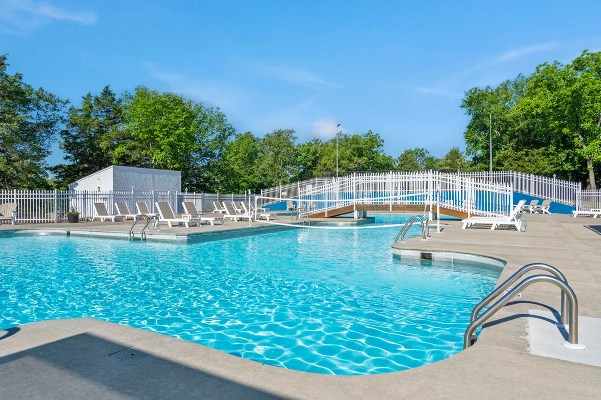 An inviting outdoor swimming pool surrounded by lounge chairs and a picturesque wooden bridge, all set within lush greenery. The area is bathed in the soft light of either early morning or evening, suggesting a peaceful, relaxing environment for couples to unwind together.