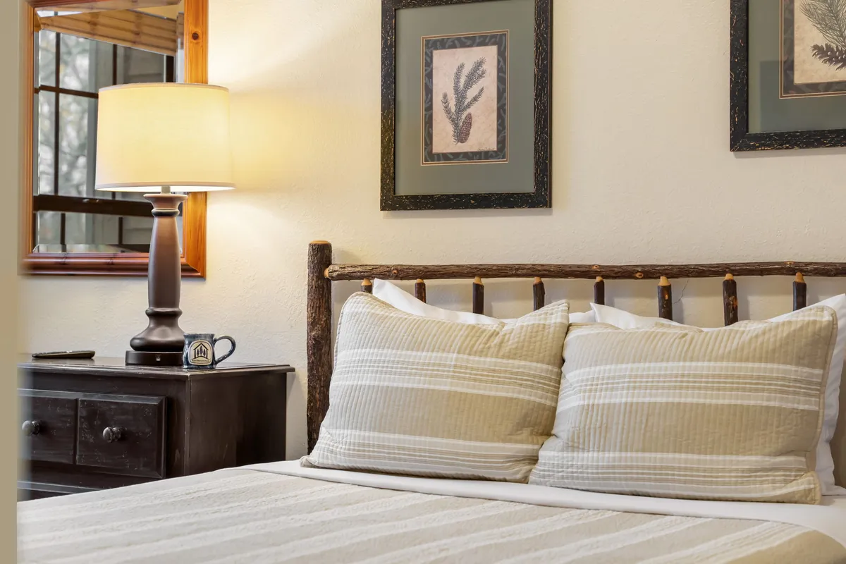 A softly sunlit queen bed with striped pillows, a lamp on the nightstand, and framed artwork above. The bedding appears plush and inviting, while sunlight filters through a window, creating a calm and restful atmosphere.